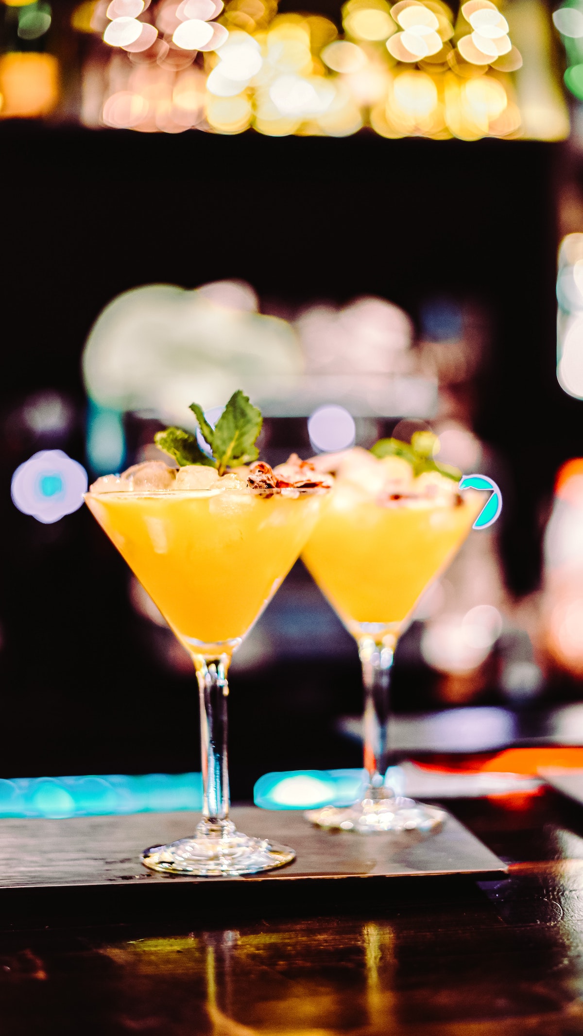 2 cócteles amarillos en vasos de martini adornados con hierbas encima de un bar.